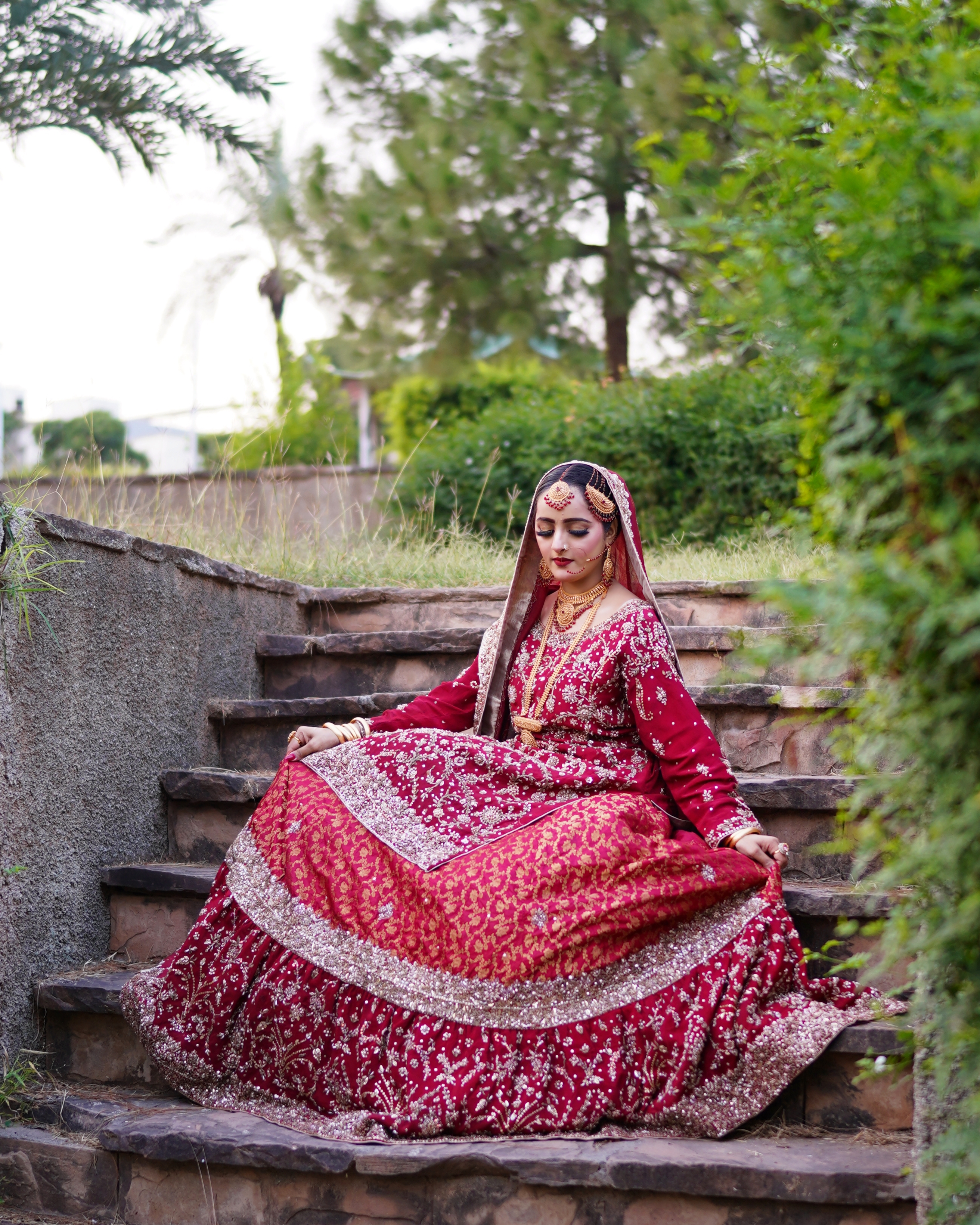 Pakistani bride sitting on garden stairs wearing a red embroidered bridal lehenga and traditional gold jewelry during an outdoor wedding photoshoot by AM Productions.