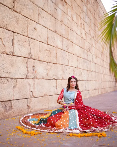 A Pakistani Mehndi bride sitting gracefully on the floor with her multi-colored paneled lehenga beautifully spread around her in a circle against a giant rustic stone block wall, c