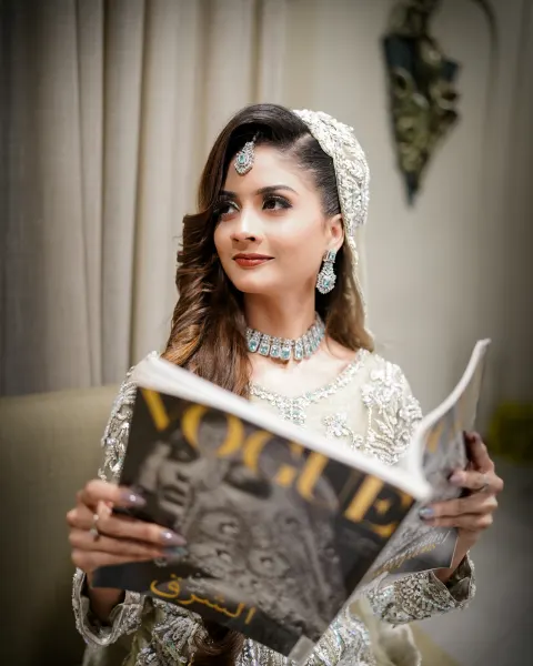A glamorous Pakistani bride looking away while holding a Vogue magazine, wearing an ivory white bridal dress with silver embellishments and diamond jewelry, captured by AM Producti