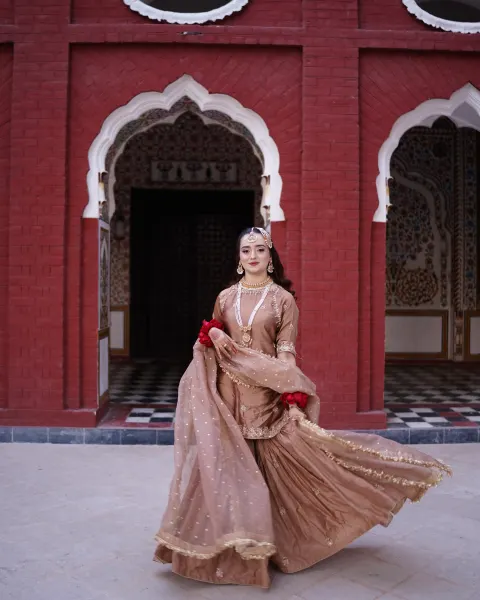 A Pakistani bride gracefully twirling her metallic mocha-brown gharara in front of a grand red brick heritage building with traditional Mughal arches, captured by AM Productions.