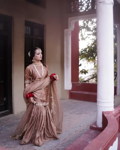A stunning Pakistani bride posing candidly on a stone veranda of a heritage building, wearing a metallic mocha-brown silk gharara with fresh red rose gajras, shot by AM Productions