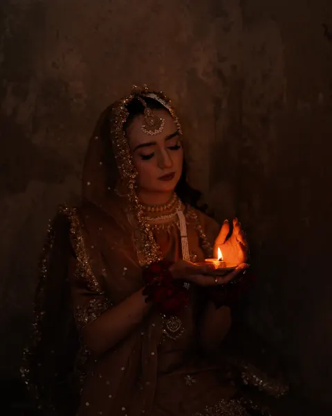 A close-up portrait of a Pakistani bride with closed eyes, gently holding a lit clay lamp (diya) that illuminates her face in a dark, moody setting, captured by AM Productions.