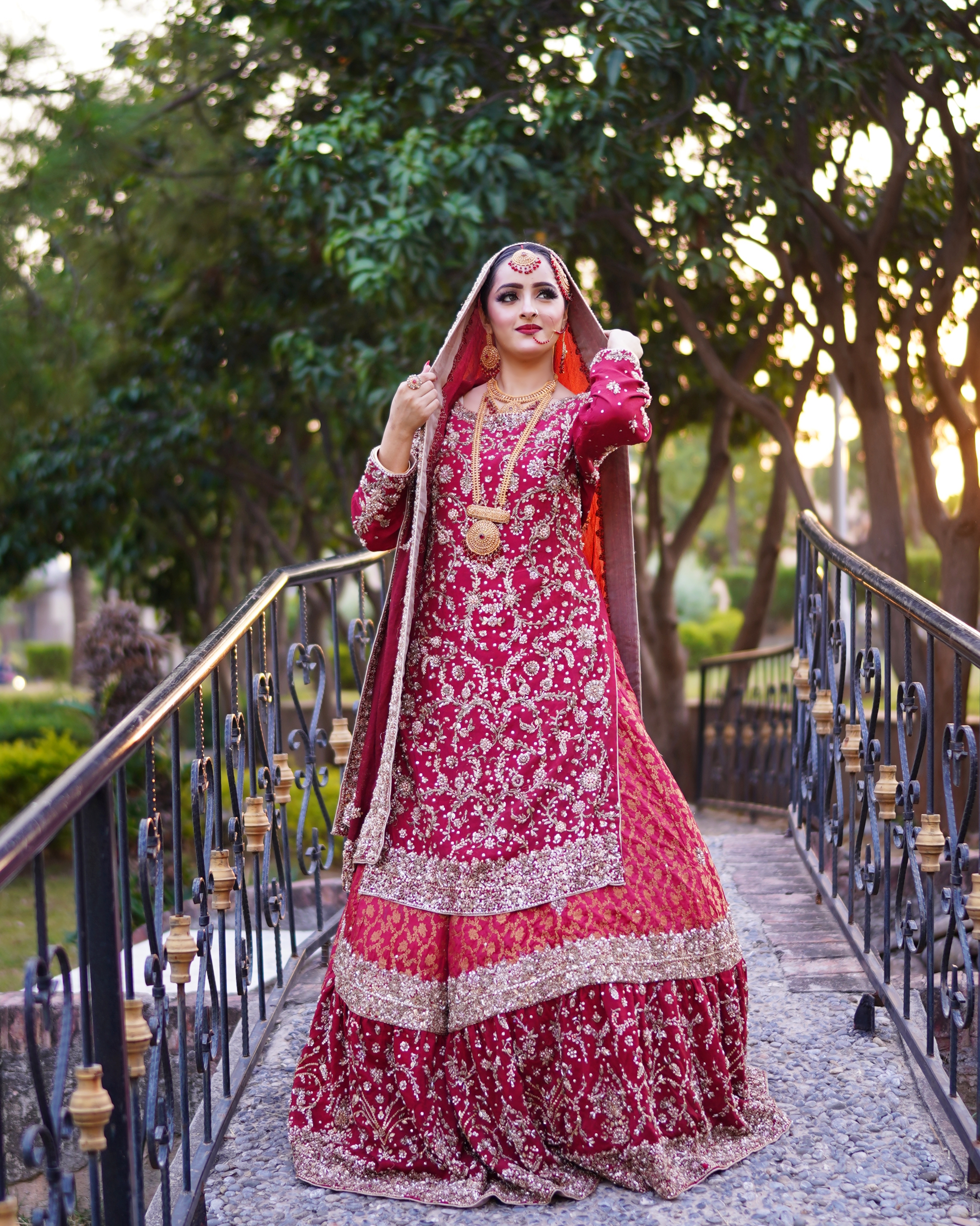 A Pakistani bride in a heavily embroidered crimson red and maroon lehenga kurti set posing on a stone bridge with iron railings, captured by AM Productions.