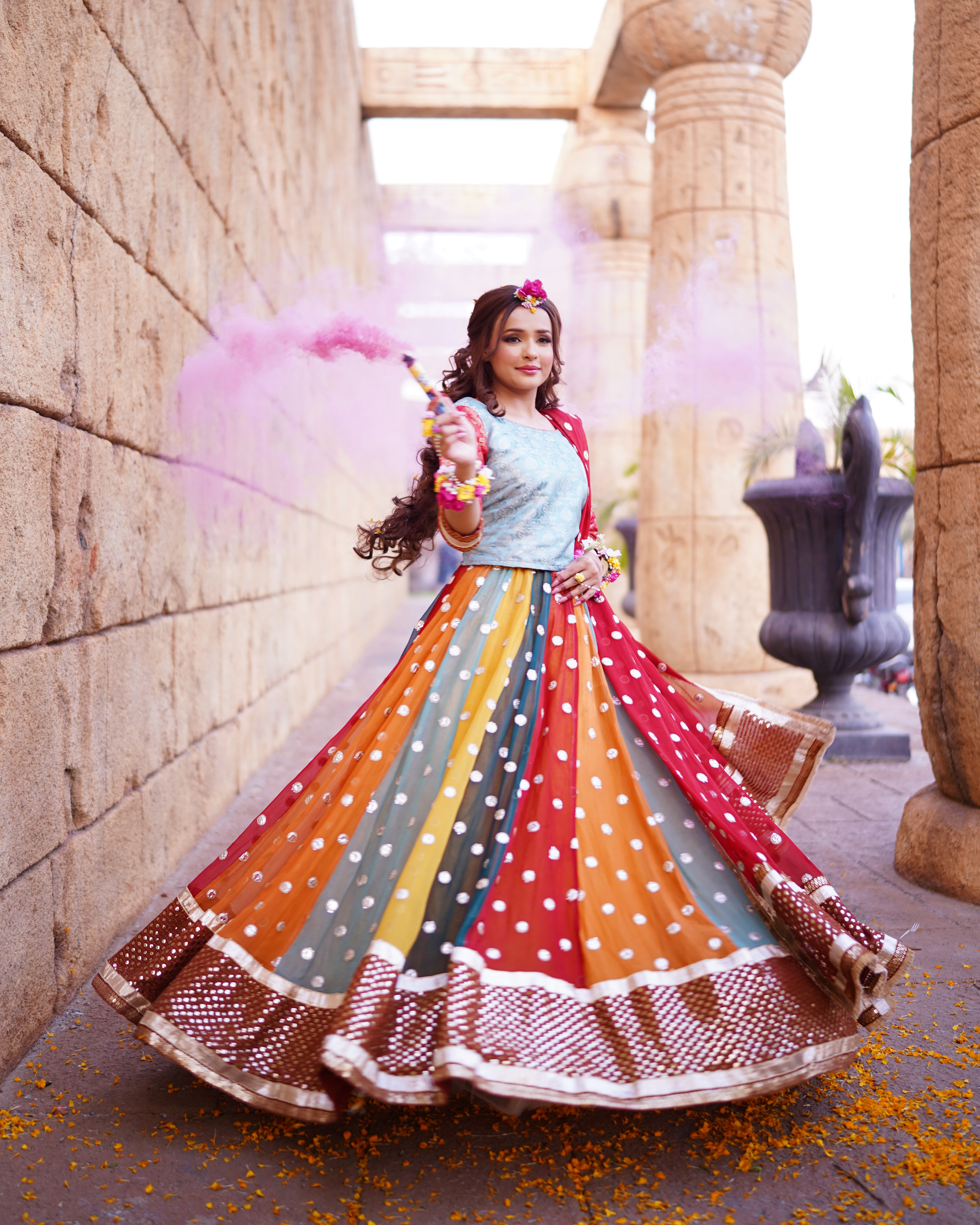 A Pakistani Mehndi bride twirling in a colorful multi-paneled lehenga and light blue choli while holding a pink smoke bomb near an ancient Egyptian-themed stone wall, shot by AM Pr