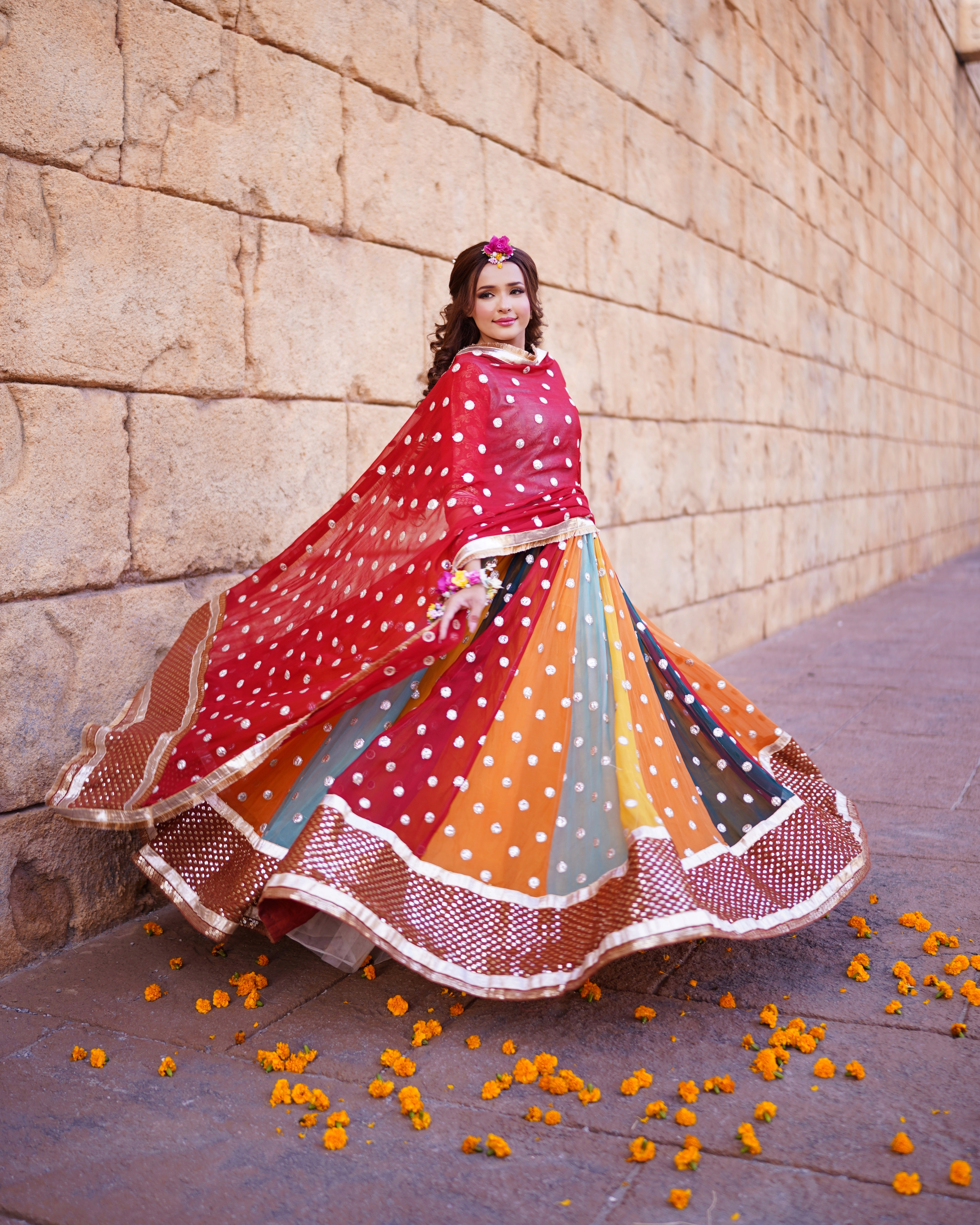 A beautiful Pakistani Mehndi bride looking over her shoulder while twirling in a colorful paneled lehenga and a red chunri-style dupatta against a large sand-colored stone block wa