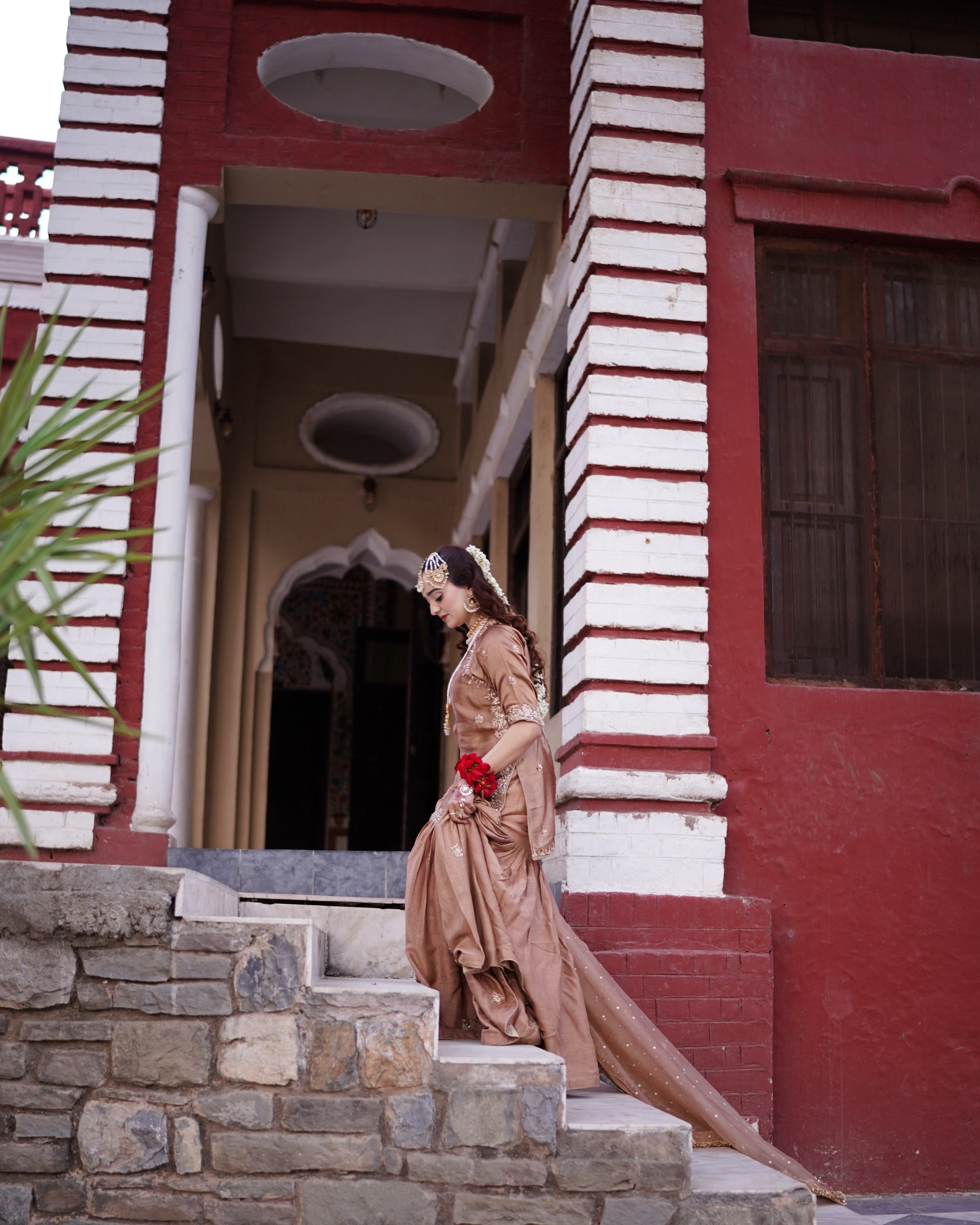 A Pakistani bride walking down rustic stone steps outside a red and white colonial-style heritage building while looking down, captured by AM Productions.