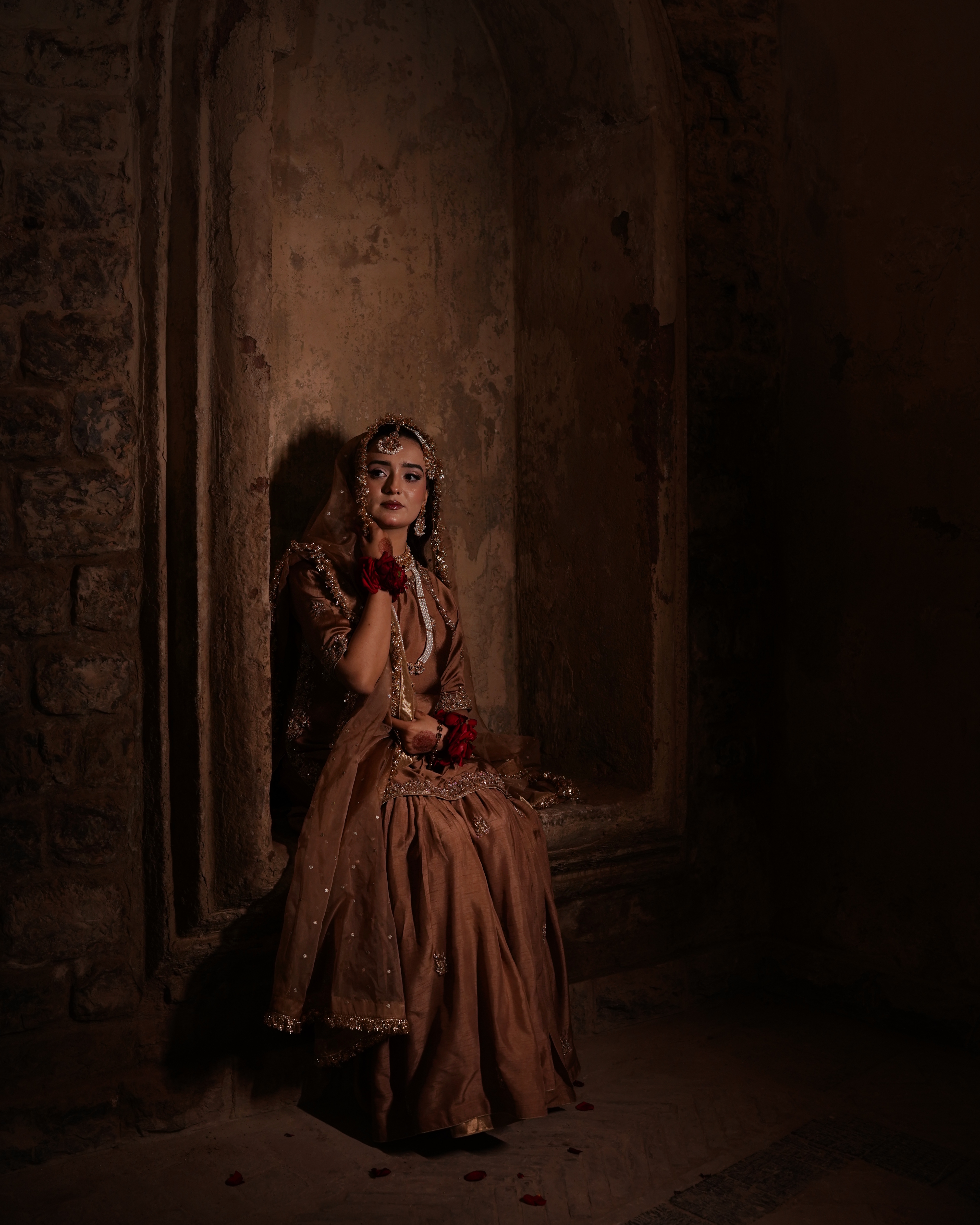A solo Pakistani bride sitting in a dimly lit rustic stone archway alcove, wearing a metallic mocha-brown gharara with deep red roses on her wrists, featured in a moody wedding ree